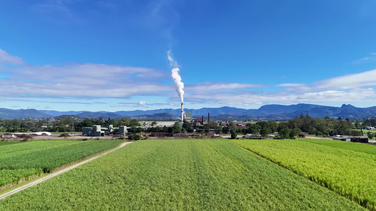Drone footage captures expansive green fields with a distant factory emitting smoke under clear blue skies