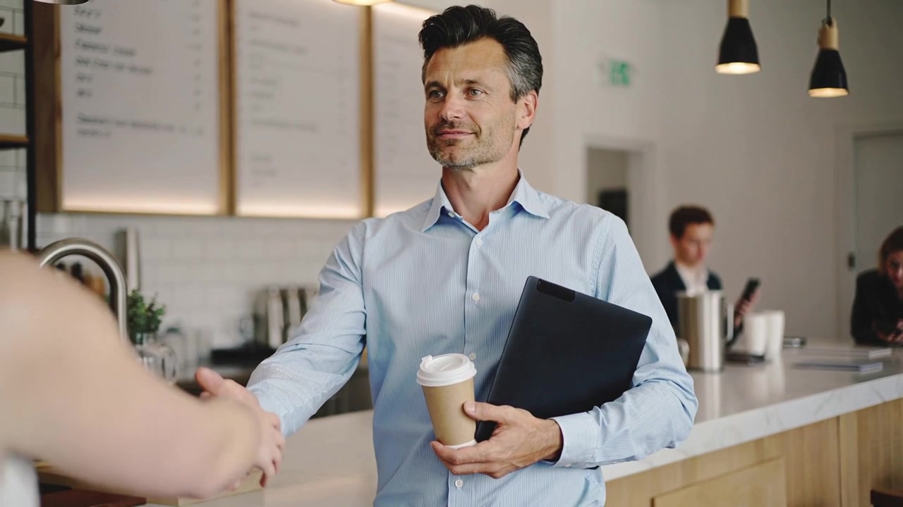 Un uomo in camicia blu, con un laptop al bancone di un caffè, si allunga per stringere la mano a un collega che sta entrando.