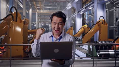 asian male professional worker standing with his laptop with industrial robotic arms working, he raises his fist up with screaming goal