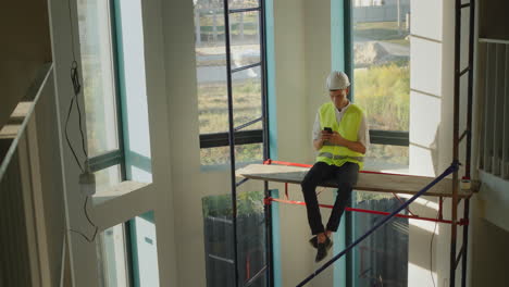 A-worker-uses-a-smartphone,-sits-high-on-scaffolding-inside-the-house