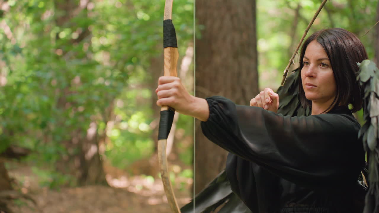 Huntress with black wings aims arrow in lush forest, standing alert and determined, symbolizing precision, strength, and mystic warrior energy in tranquil natural light surrounded by green trees