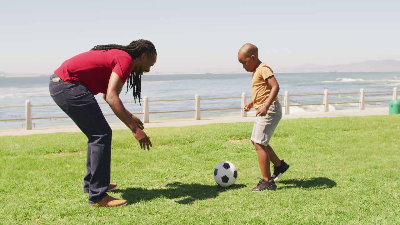 Video of happy african american father and son playing soccer outdoors and having fun