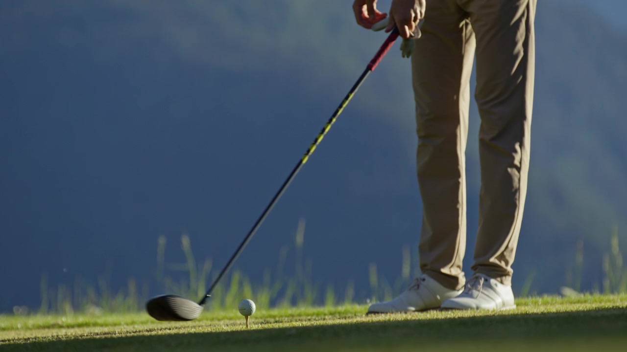 Inquadratura dal basso di un golfista che esegue uno swing di prova accanto a una pallina da golf posizionata sul tee su un campo alpino a Verbier, Svizzera. Allenamento, preparazione e tecnica di golf nelle Alpi svizzere
