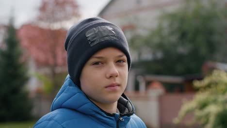 close-up of kid in black beanie and blue jacket outdoors, with calm expression and slight frown, surrounded by vibrant autumn trees and a blurred background featuring red-roofed house and greenery