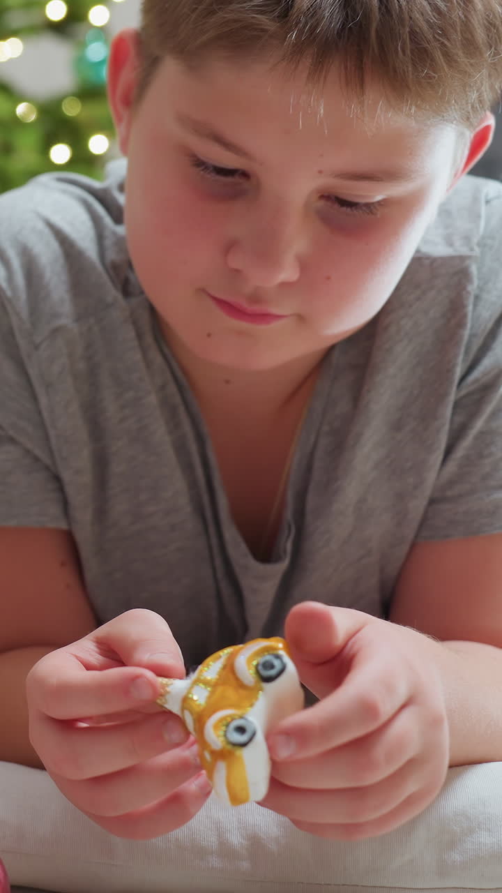 Ragazzo che esamina una pallina di Natale crepata, luci dell'albero con bokeh delicato, espressione pensierosa, mani che tengono una pallina a strisce dorate, cuscino in soggiorno, pallina rosa sparsa sul pavimento, luce naturale, silenzio