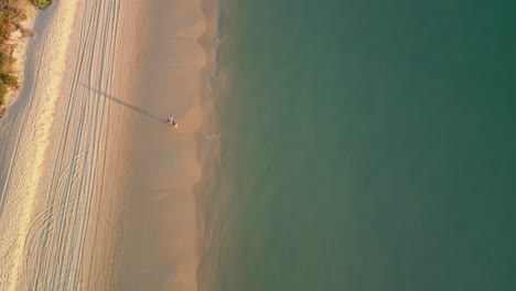 vertical drone follows white sand beach path, sea water coastline, couple walks along shore, aerial tops down