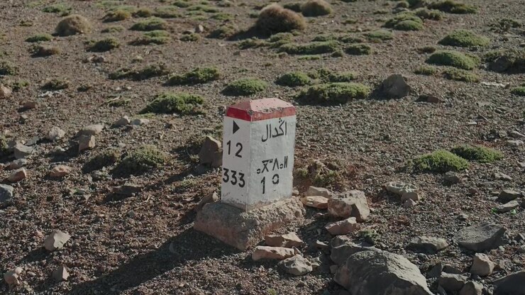 Rød og hvid vejmilepæl, der markerer Agdal, blandt sparsom mos og klipper på et Høj Atlas-plateau i Marokko.