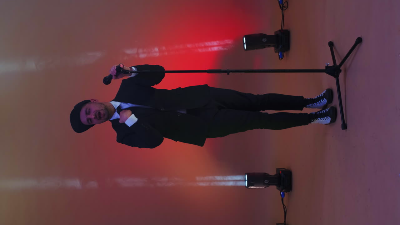 Vertical video. A funny man sings and dances into a microphone on a stand in the neon light of lamps and in a Studio on a colorful bright background