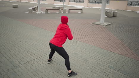 woman performing outdoor winter workout, stretching into a side lunge on paved ground, wearing red hoodie and black leggings, in cold weather environment surrounded by urban architecture