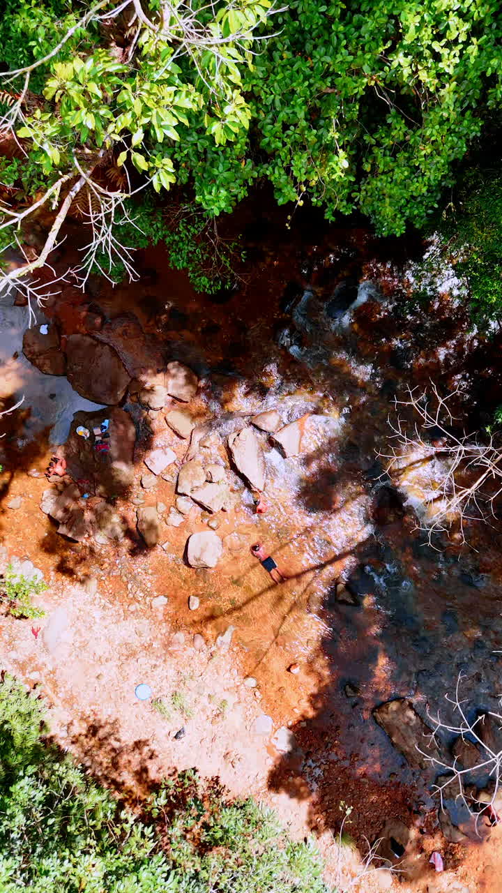 Descending above the shallow river flowing by the rocky landscape. Man lies relaxed in the clear water. Vertical video.