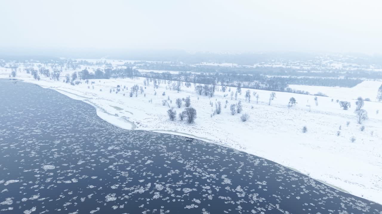 Drone footage capturing a serene winter landscape with a snow-covered shoreline and a partially frozen river. The aerial perspective offers a tranquil view of nature's cold beauty during snowfall