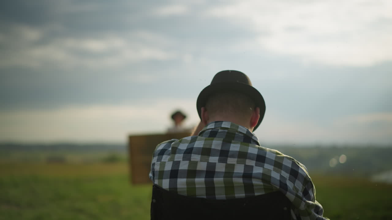 검은 모자를 입은 남자가 잔디에 앉아있는 동안 그것을 조정하는 근접적인 뒷면. 모기는 배경에 흐릿한 여자가 그림을 그리면서 그의 주위에 날아갑니다.