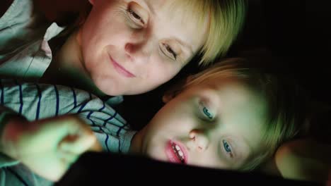 Glückliche-Mutter-Mit-Ihrer-Tochter-Zusammen-Spiele-Ich-Auf-Dem-Tablet-Liege-Nebeneinander-Im-Bett