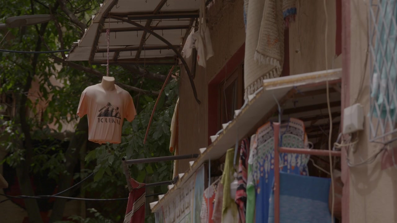 A souvenir t-shirt hangs suspended alongside patterned woven textiles outside a rustic roadside artisan shop, showcasing local village commerce and tourism in Morocco’s High Atlas Mountains region.