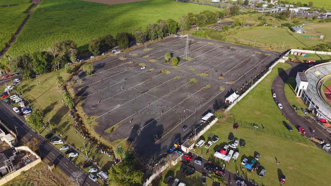 Push in drone shot of driving or racing event in a Mauritius car park. A car speeds past a large white event tent and spectators, capturing the grassroots motorsports action