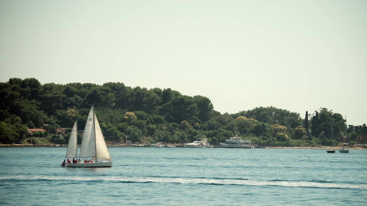 Barca a vela che trasporta persone in una crociera estiva attraverso le vaste acque blu dell'oceano, passando una costa verde lussureggiante con barche ormeggiate.