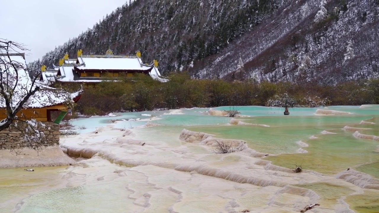 En svepande panoramabild som stiger från närbilder av gula travertinterrassväggar för att visa de vidsträckta turkosa poolerna och ett snötäckt tempelkomplex i Huanglongs natursköna område, Kina.