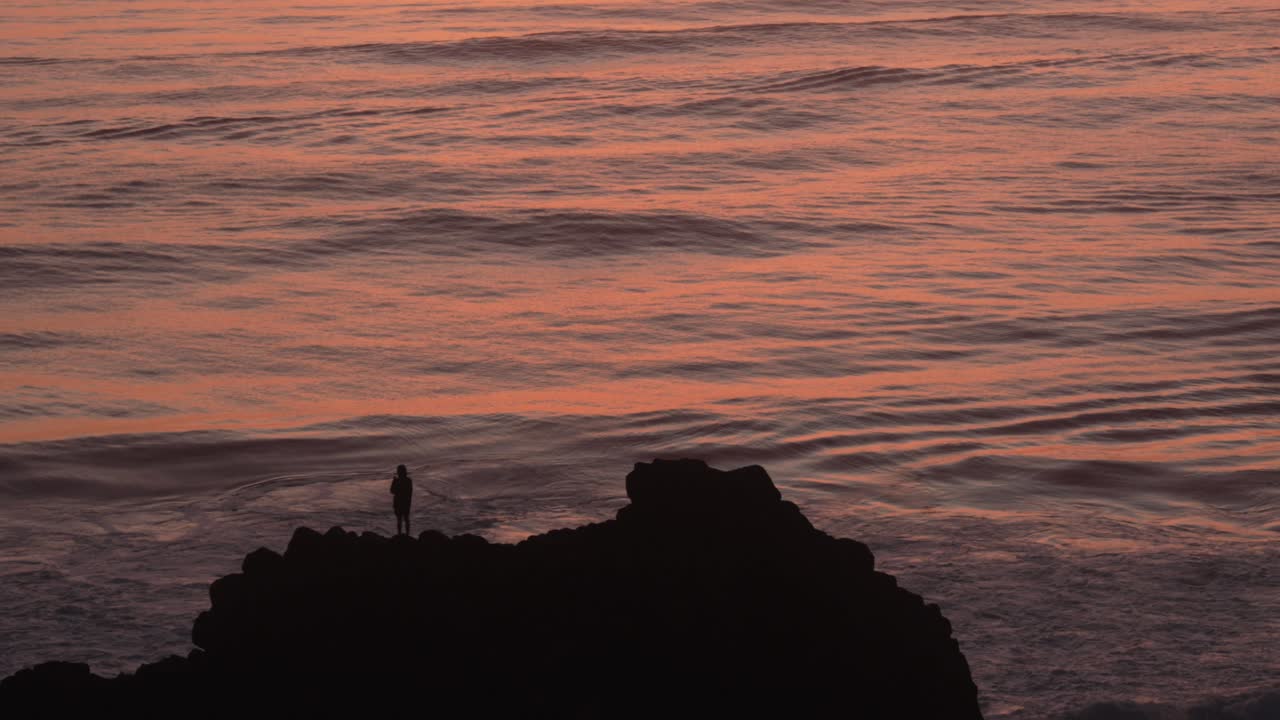 Person standing on rock at the coastline of Coolangatta silhouette shot during sunrise, orange reflection waves.