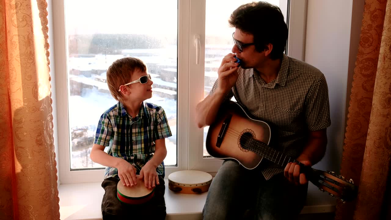 papá está tocando la guitarra y el hijo está tocando el tambor sentado en el alféizar de la ventana