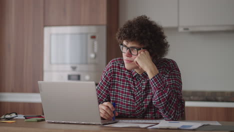 Estudiante-Masculino-De-Pelo-Rizado,-Un-Joven-Atractivo-Con-Gafas-Está-Estudiando-En-Casa-Usando-Una-Computadora-Portátil-Escribiendo-En-Un-Cuaderno.-Estudiante-Universitario-Usando-Una-Computadora-Portátil-Viendo-Un-Seminario-De-Aprendizaje-En-Línea-A-Distancia