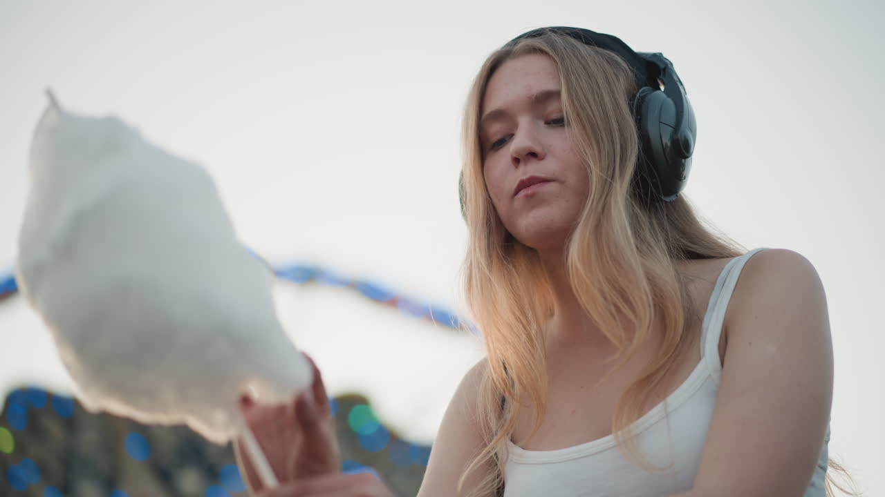 DJ caucasico con cuffie, zucchero filato da vicino al tramonto al luna park, espressione contemplativa, calda retroilluminazione, bokeh colorato dell'attrazione, mano che cerca la golosità soffice, toni tenui cinematografici, intimo