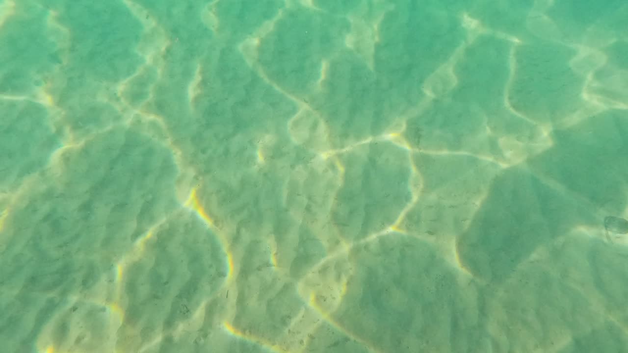 Clear turquoise water reveals a sandy sea floor with moving sunlight patterns. Gentle underwater camera movement creates dynamic, shimmering textures in bright natural light