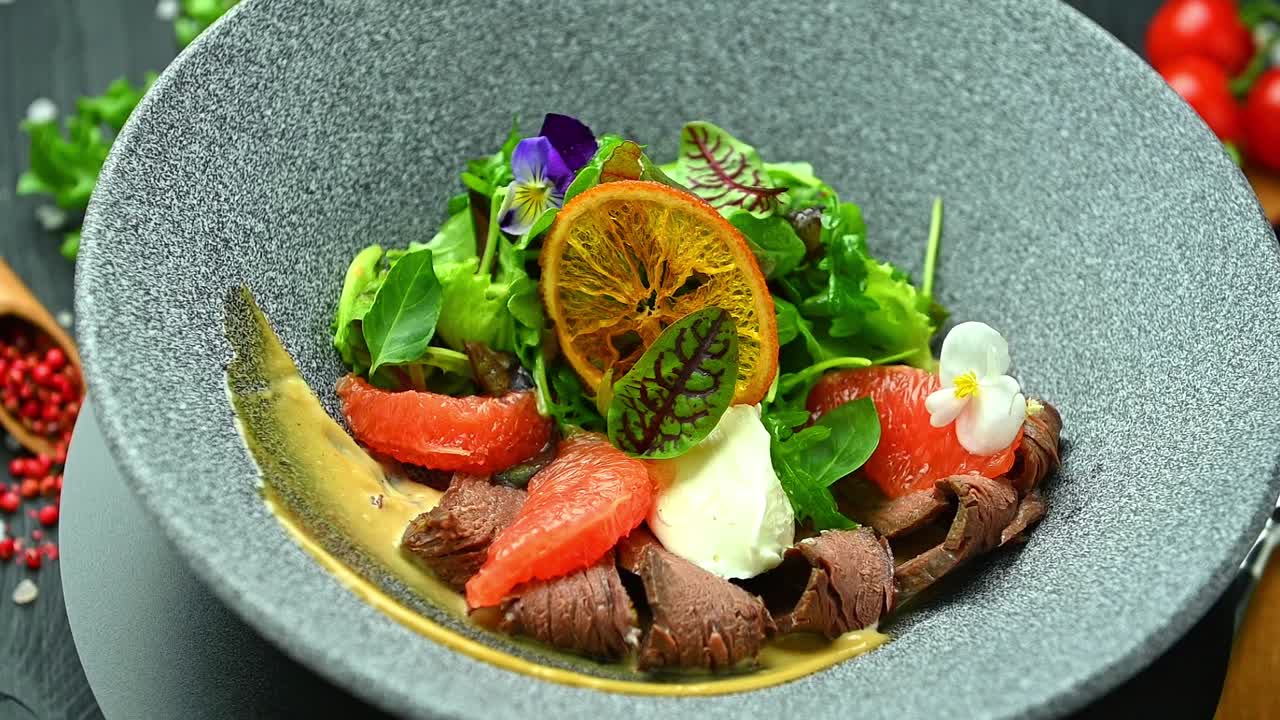 Frisk salat med oksekød og citrusfrugter serveret i en skål på et sort bord. En skål indeholder en frisk salat med skiver af oksekød, grapefrugt og krydderurter på et sort bord.