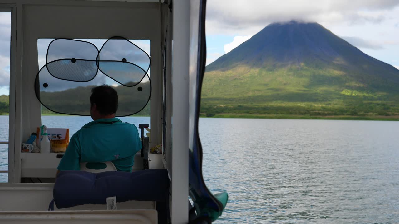 Front shot, inside a moving boat a man driving the boat on Lake Arenal in Costa Rica, a scenic view of Arenal volcano in the background.