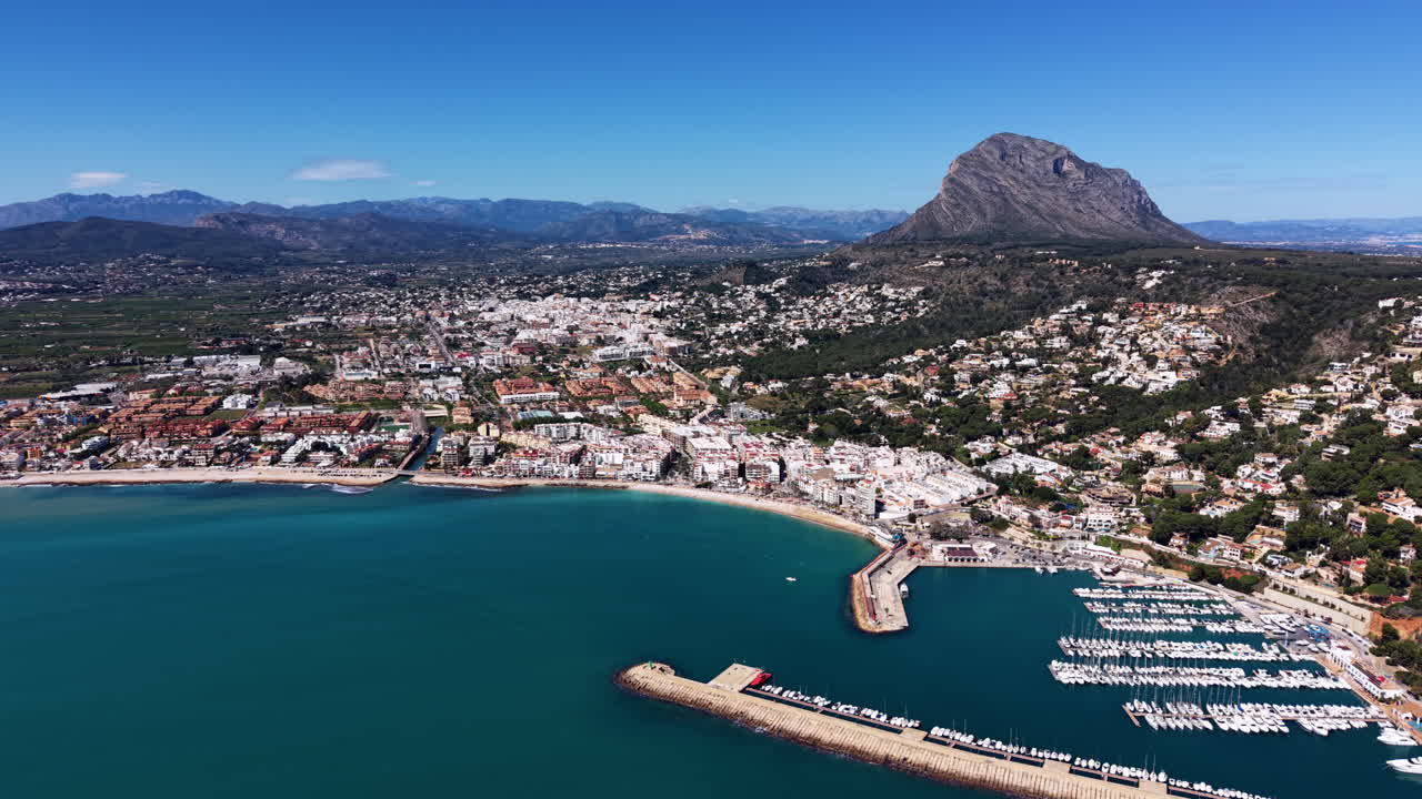 Cittadina costiera di Javea sulla Costa Blanca, Spagna, con baia mediterranea turchese, porto turistico con barche ormeggiate, edifici bianchi e suggestivo sfondo montuoso roccioso