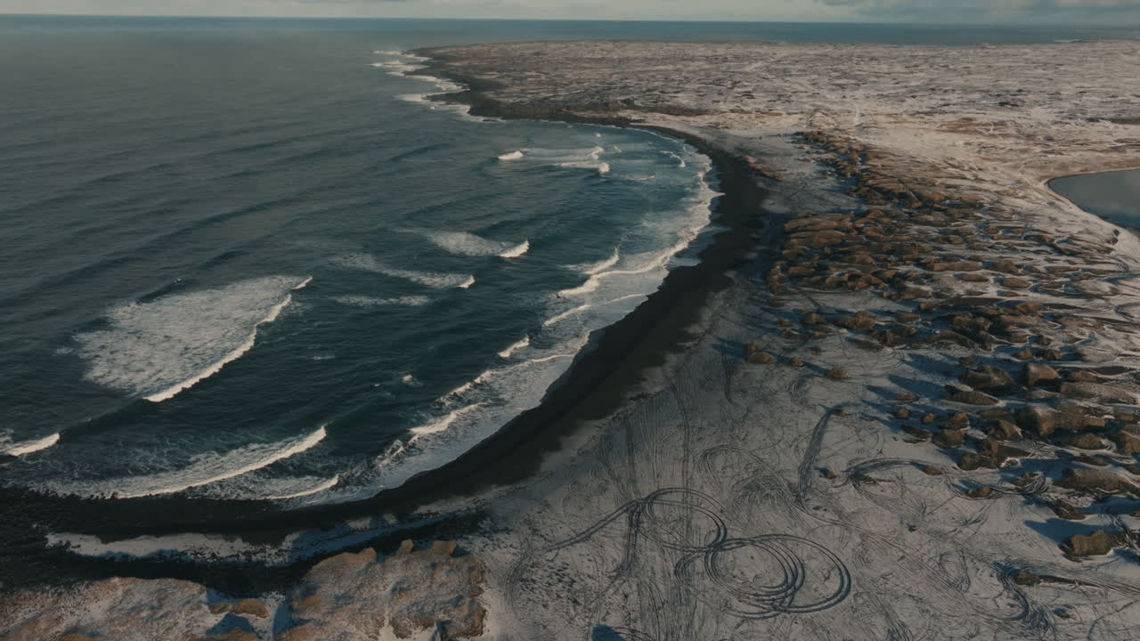 Drone aerial view of a snow-covered volcanic coastline in Iceland with a black sand beach, ocean waves, and tire tracks visible on the snowy terrain. Winter coastal landscape and remote Nordic wildern