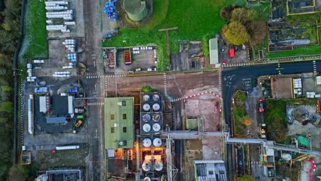 Forward-moving-overhead-aerial-footage-of-a-large-industrial-plant-showing-pipework-structures,-buildings,-cooling-towers,-steam,-and-work-vehicles