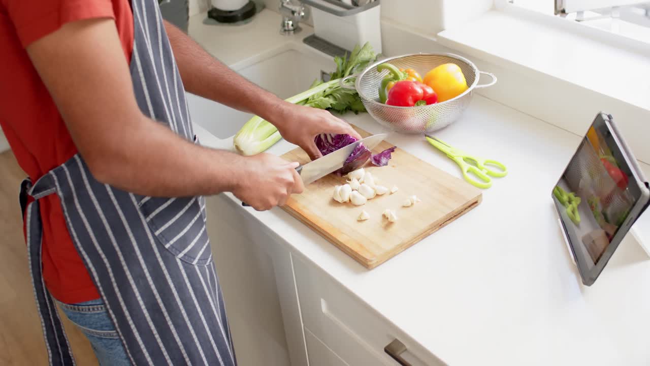 sección media de un hombre biracial en delantal cortando verduras y usando tableta en la cocina, cámara lenta