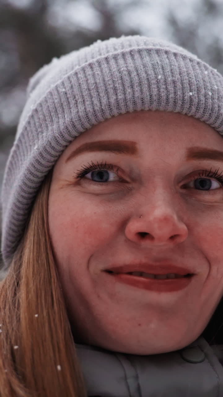 Extreme close-up of woman's face in gray knit beanie smiling softly with snowflakes on eyelashes and hair, showing emotion, detail, and winter atmosphere in forest with blurred background and cold light