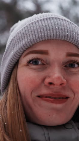 extreme close-up of woman's face in gray knit beanie smiling softly with snowflakes on eyelashes and hair, showing emotion, detail, and winter atmosphere in forest with blurred background and cold light