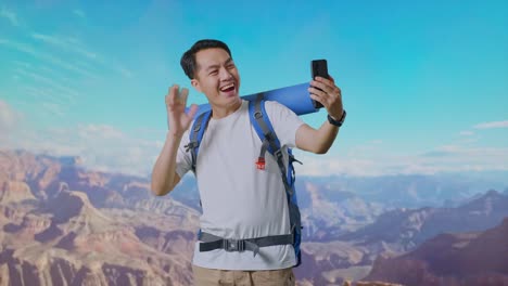 asian male hiker with mountaineering backpack waving his hand and pointing back while having a video call on smartphone while traveling at the top of mountain