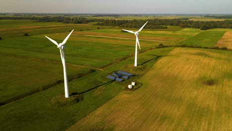 drone shot of two working wind turbines and a few solar panels producing green electric energy on a cultivated field on a sunny summer day, use of renewable resources of energy, zoom out