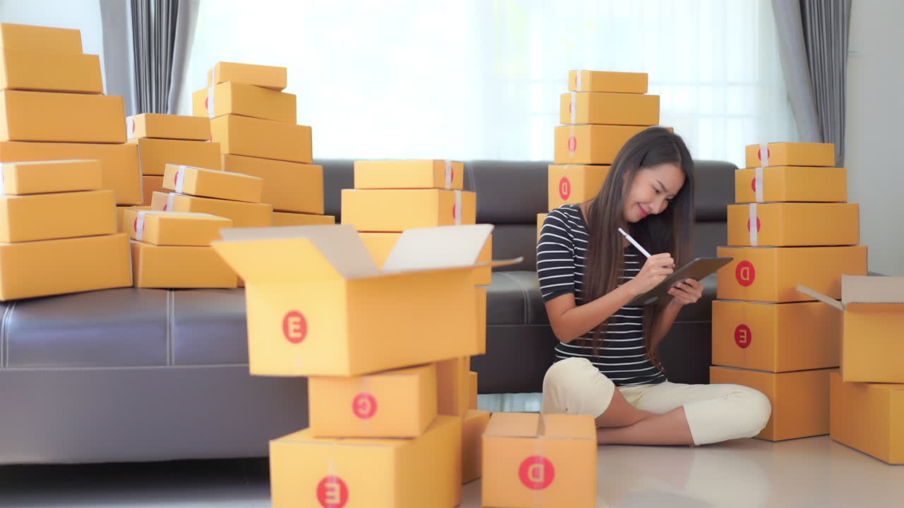 Static shot of a cute Asian lady sitting on the floor surrounded by boxes while writing something on a tablet and smiling