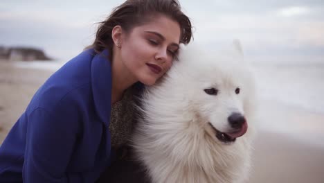 mujer joven sentada en la arena y abrazando a su perro de la raza samoyed por el mar. mascota peluda blanca en la playa divirtiéndose