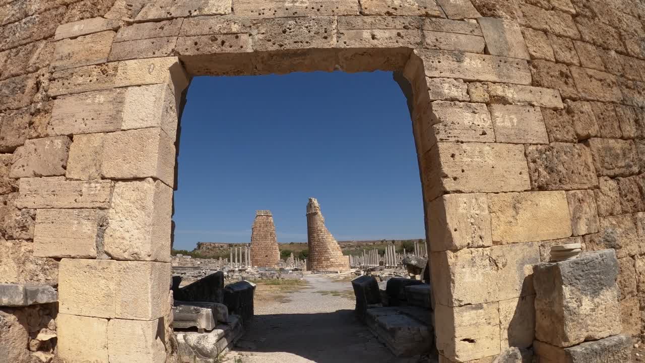 Aerial view of ancient stone archway leading toward twin pillars within historic Patara ruins in Antalya Province, Turkey, surrounded by weathered masonry and open landscape