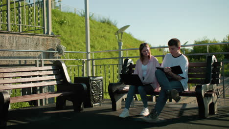 dama de ropa rosa sentada al aire libre con su amiga, ambas estudiando juntas, quita la mano de la tableta del niño y continúa trabajando en su portátil, el fondo presenta colina verde y postes de luz