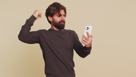 happy excited joyful man using smartphone celebrating success winning game on beige background