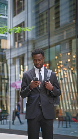 Vertical-Video-Shot-Of-Young-Businessman-Wearing-Suit-Using-Mobile-Phone-Standing-Outside-Offices-In-The-Financial-District-Of-The-City-Of-London-UK-Shot-In-Real-Time-1