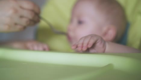 mom-feeds-baby-in-green-highchair-focus-on-little-boy-hand