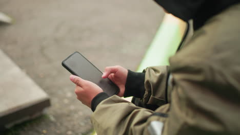 close up side view of kid in jacket holding smartphone with both hands while sitting outdoors, deeply focused on operating digital device