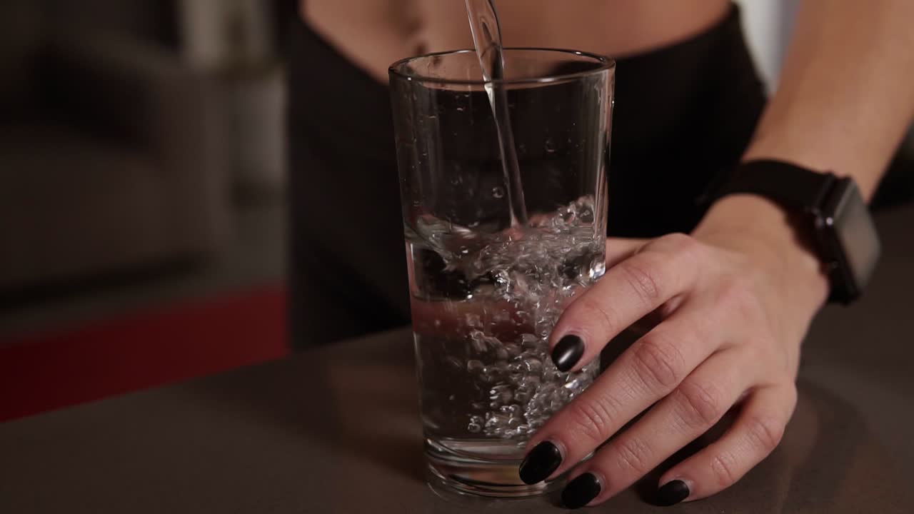 cerca de las manos de la mujer con manicura oscura vierte el agua en el vaso de pie en la mesa de la cocina. alimentación saludable, agua después del entrenamiento