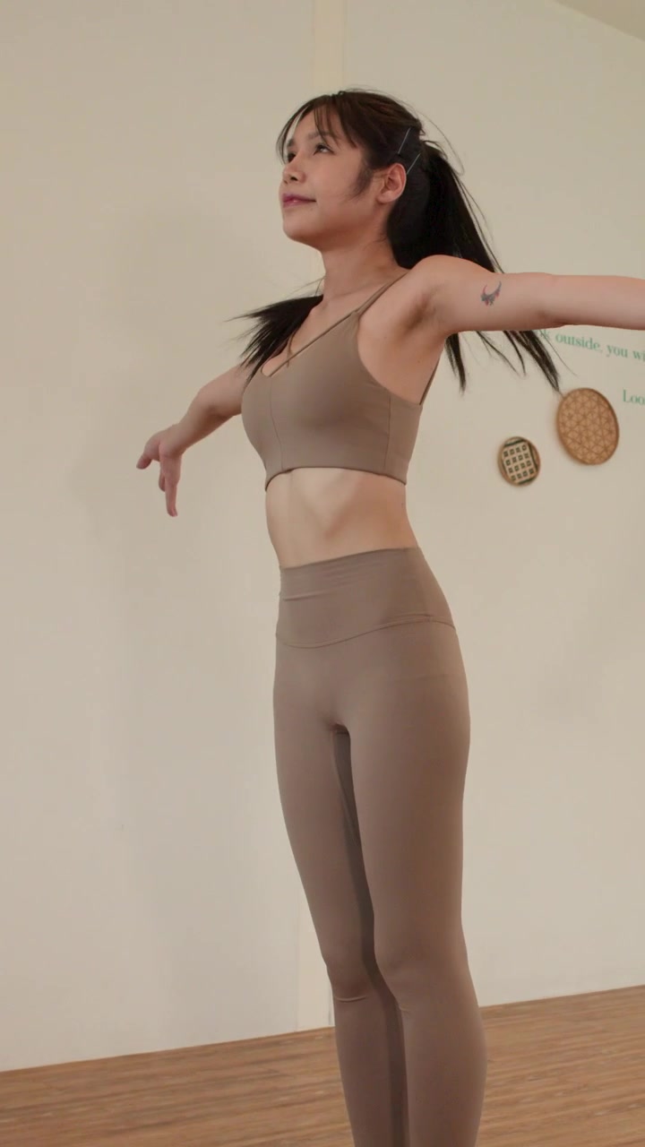 An Asian woman smoothly transitions from a standing forward fold pose in a studio.