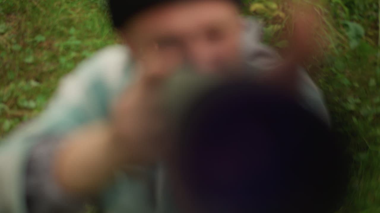 La vista del objetivo cenital muestra a un hombre apuntando, silueta de barril vertical con una pequeña apertura central, manos y torso borrosos, suelo cubierto de hierba y hojas dispersas visibles, los roles cambian de fotógrafo a excursionista.