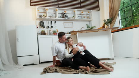married couple and a newborn sit on the plaid on the floor in the in kitchen. happy family portrait.