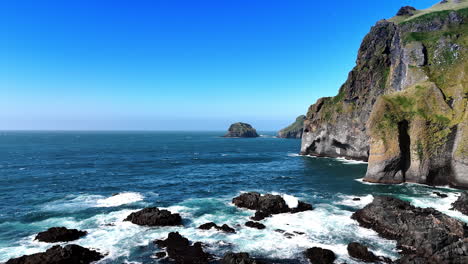 black rock stick out of water at the shore. white waves splash by the rocks. steep mount cliff towering over the waterscape.
