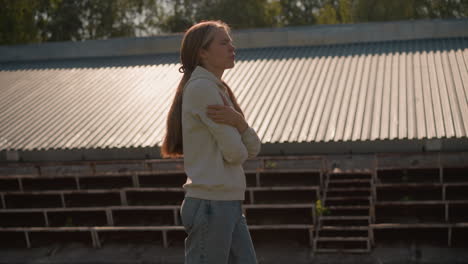 young girl with long hair tied back in hoodie and jeans walks by empty stadium bleachers on sunny day, arms folded, appearing reflective and thoughtful, surrounded by quiet, empty seating